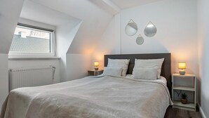 Un dormitorio con una cama grande, dos mesitas de noche con lámparas, un radiador, una ventana y una pequeña estantería con una planta.