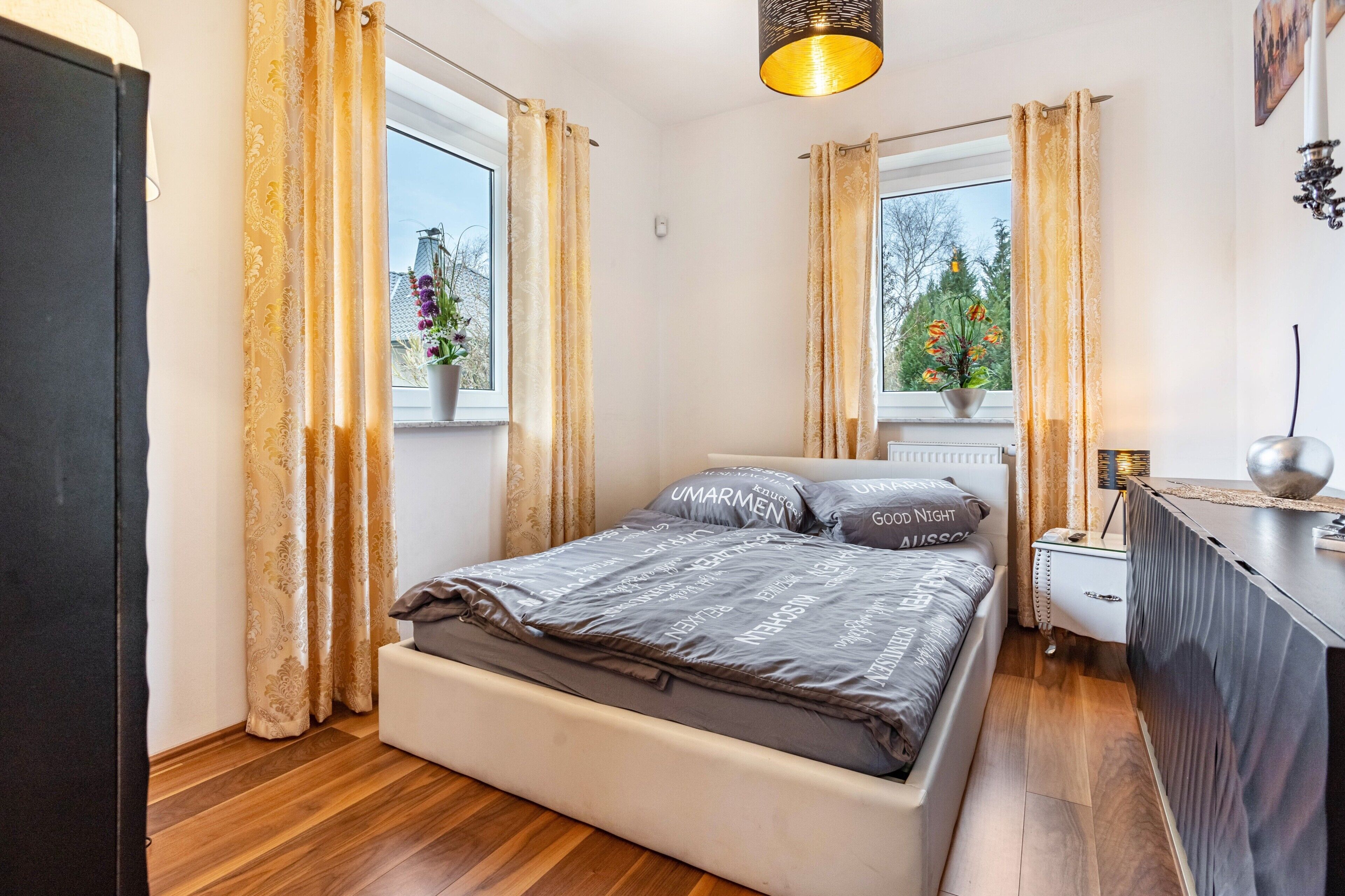 Ein Schlafzimmer mit Bett, Vorhängen, einem Nachttisch, einer Lampe und einem Fenster mit Blick auf Bäume.