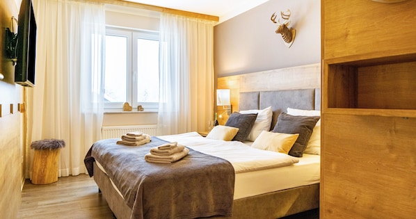 A hotel room with a bed, a television, a window with curtains, a wooden headboard, and a wall-mounted deer head decoration.