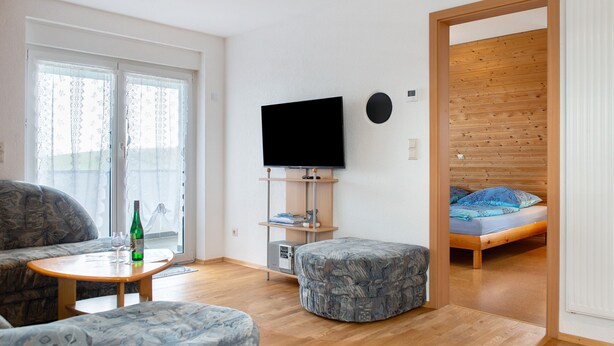 Uma sala de estar moderna com um sofá, um banquinho e uma pequena mesa com uma garrafa e copos. Uma televisão está montada na parede, e uma cama é visível através de uma porta aberta.