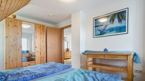 Quarto com duas camas, guarda-roupa de madeira, escrivaninha e pintura de uma rede numa praia.