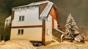 Une cabane recouverte de neige, avec une façade en bois, et un arbre également recouvert de neige à proximité.