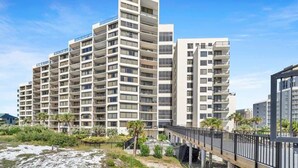 Un hôtel moderne à plusieurs étages, doté de balcons, situé près d’une plage et accessible par un pont piétonnier.