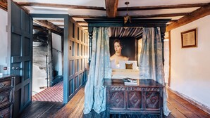 Una habitación con una cama de madera, una mesita pequeña con libros, un espejo y una ventana con cortinas.