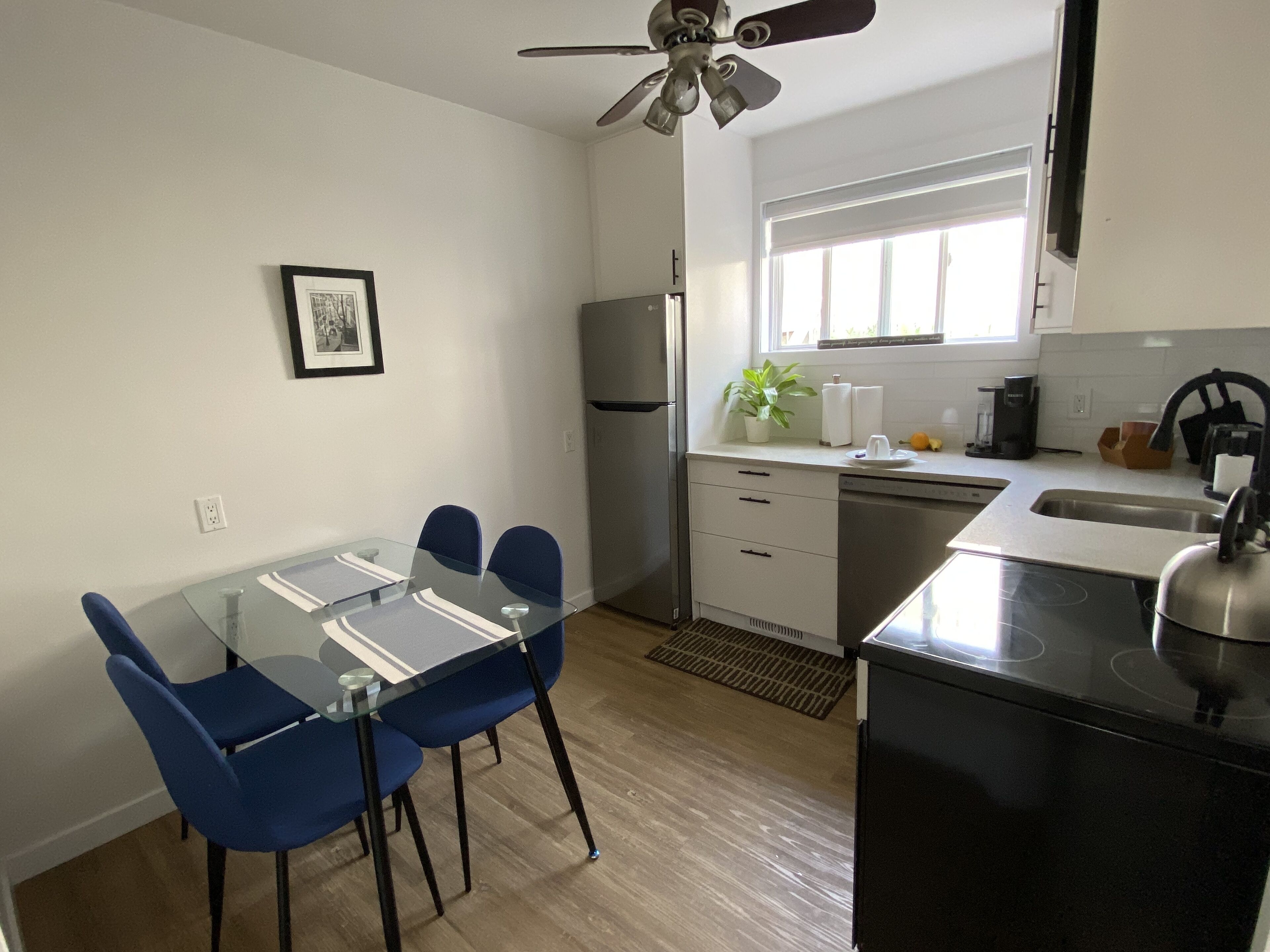 A modern kitchen with a glass dining table, blue chairs, a black stove, and a ceiling fan.