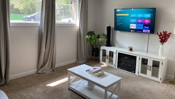 Una sala de estar moderna con un televisor de pantalla plana montado en la pared, un aparador blanco con puertas de cristal, una mesa de centro blanca con un libro y una planta en maceta.