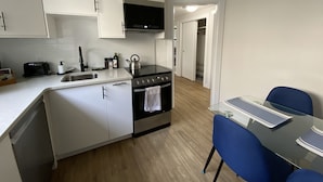 Cocina compacta con armarios blancos, estufa negra, microondas y un pequeño comedor con sillas azules.