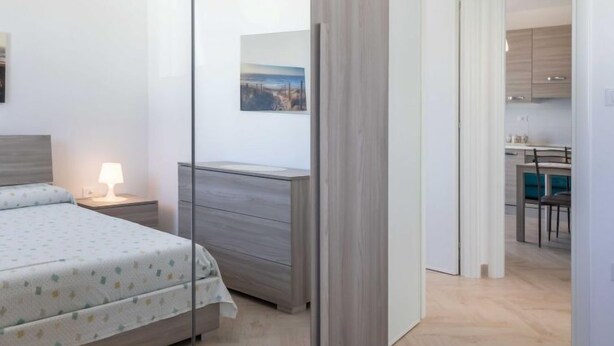 A modern bedroom with a bed, bedside table, and a dresser. There is a painting on the wall and a view into the adjacent kitchen area. - Gioia 1 by Interhome (Marina di Pescoluse)