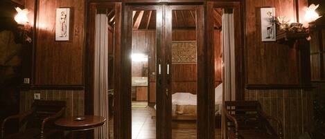 A wooden interior with a sliding door leading to a bedroom, two wooden chairs, and a small table.