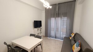 Een woonkamer met een witte tafel, twee stoelen, een bank met een kleurrijk kussen, een televisie op een dressoir en een raam met dunne gordijnen.
