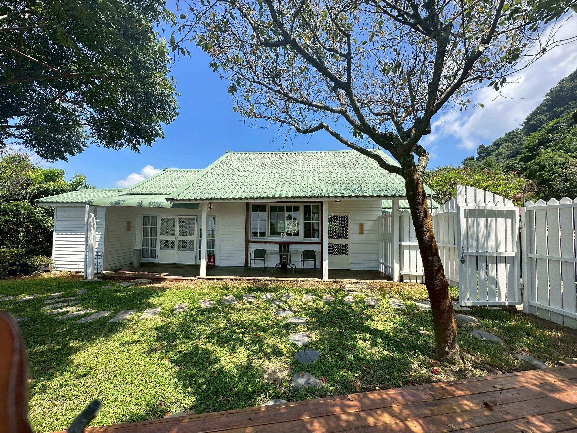 A single-story house with a green roof, a porch with two chairs, and a white picket fence.