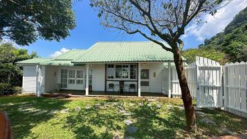 A single-story house with a green roof, a porch with two chairs, and a white picket fence.