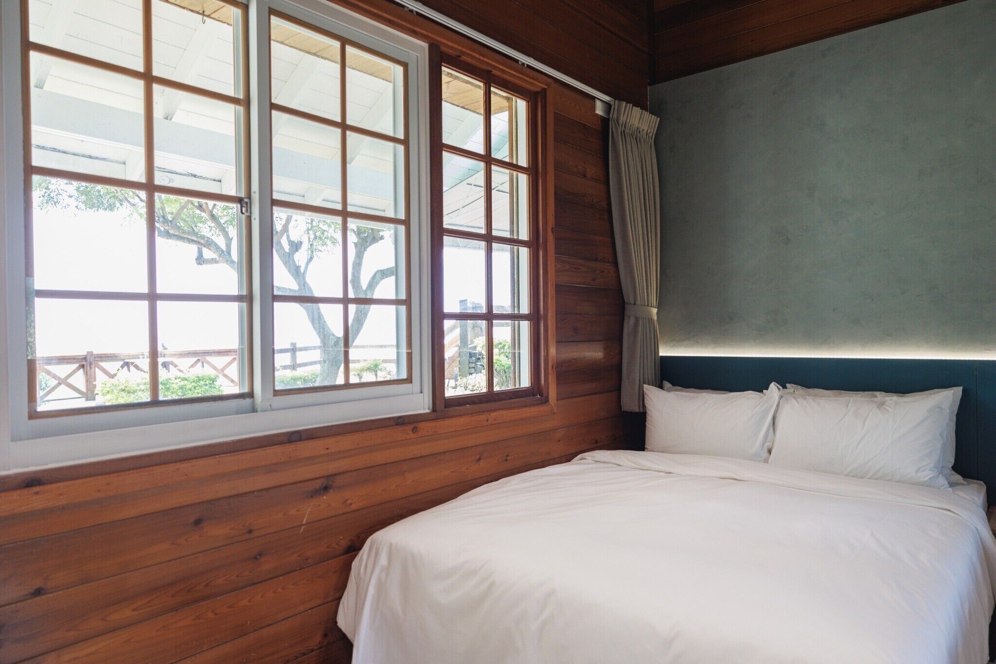 Een slaapkamer met een houten hoofdeinde, een groot raam met veel ruitjes en een bed met wit beddengoed.