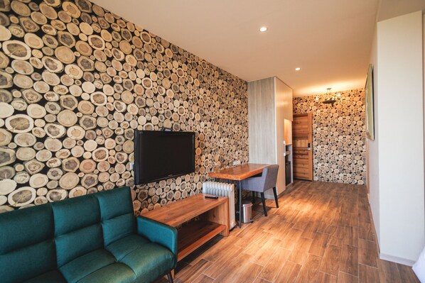 A modern living room with a wall of stacked logs, a flat-screen TV, a wooden coffee table, a teal sofa, and a small dining area with a chair and table.