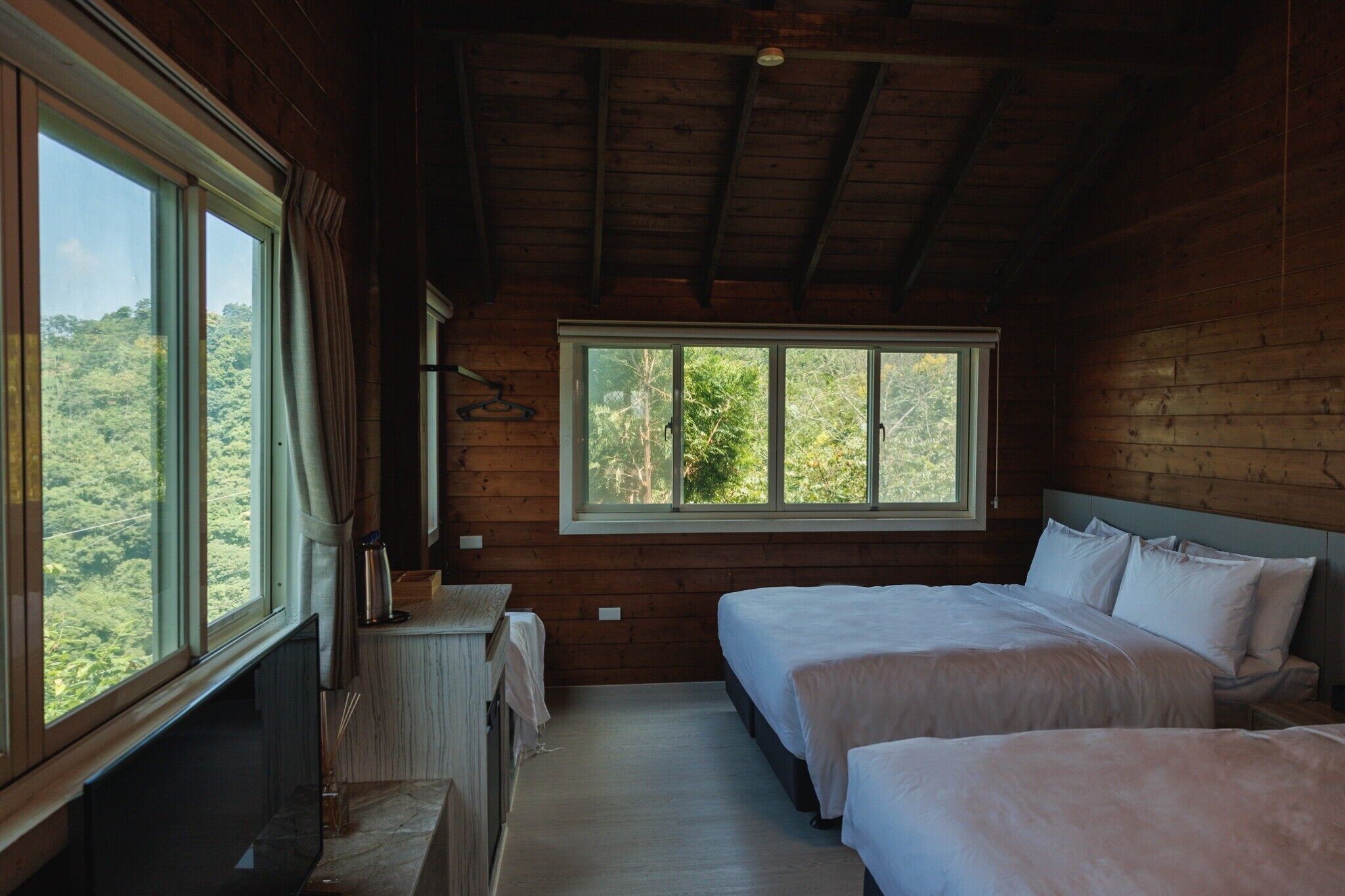 A cabin with two beds, a wooden interior, and a large window offering a forest view.