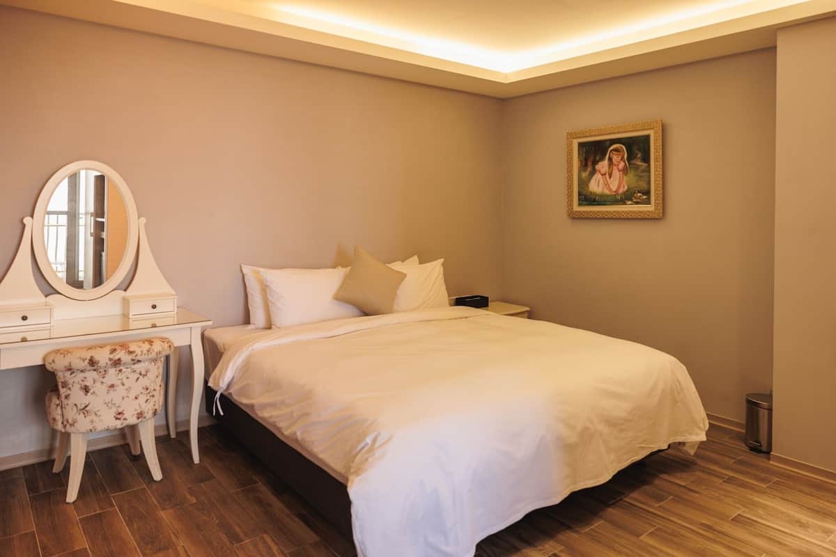 A bedroom with a bed, a vanity with a mirror, a chair, and a painting on the wall.