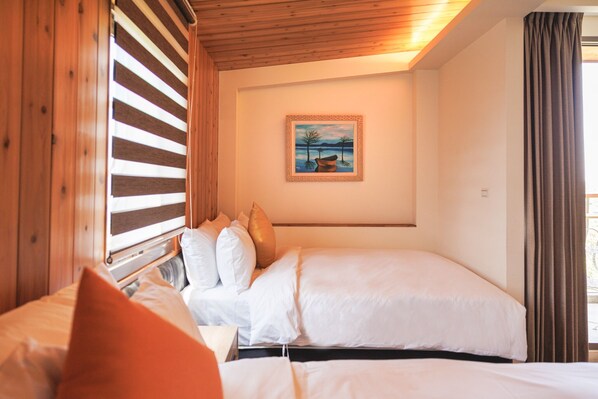 A double bed with white bedding and multiple pillows, a framed painting on the wall, and a window with blinds.