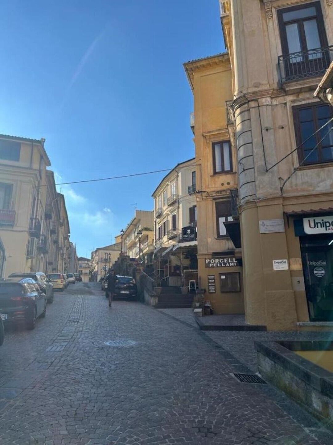 Une rue pavée bordée d’immeubles, dont un bâtiment commercial dont l’enseigne indique « UnipolSai ».