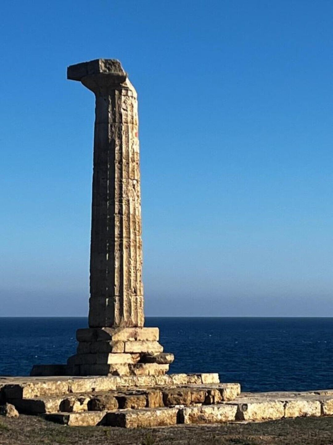 A tall, ancient stone column with a flat top, standing on a stone platform by the sea.