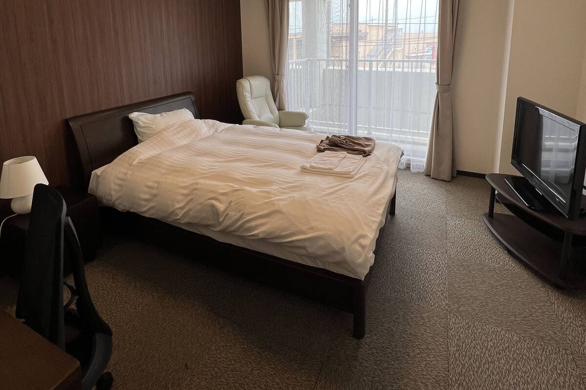 Habitación de hotel con una cama grande, una silla, un escritorio, un televisor y una ventana con cortinas.