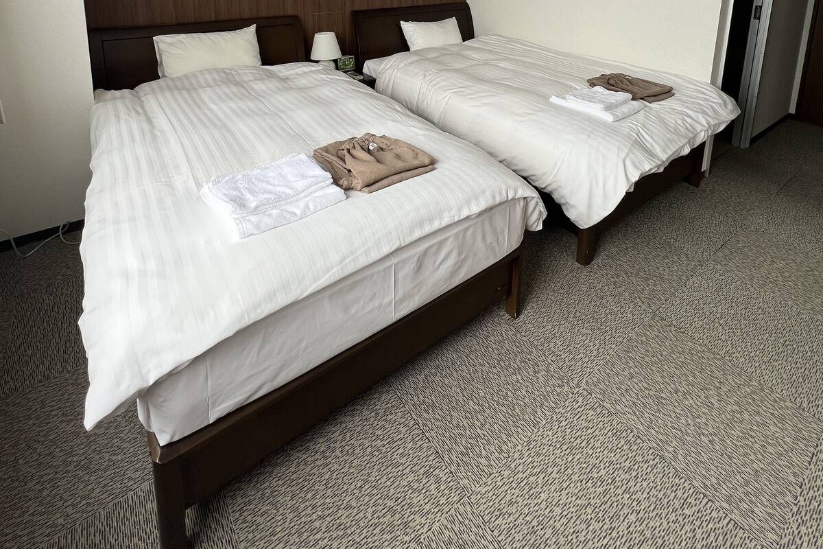 Habitación de hotel con dos camas, cabecero de madera, unidad de aire acondicionado y piso alfombrado.