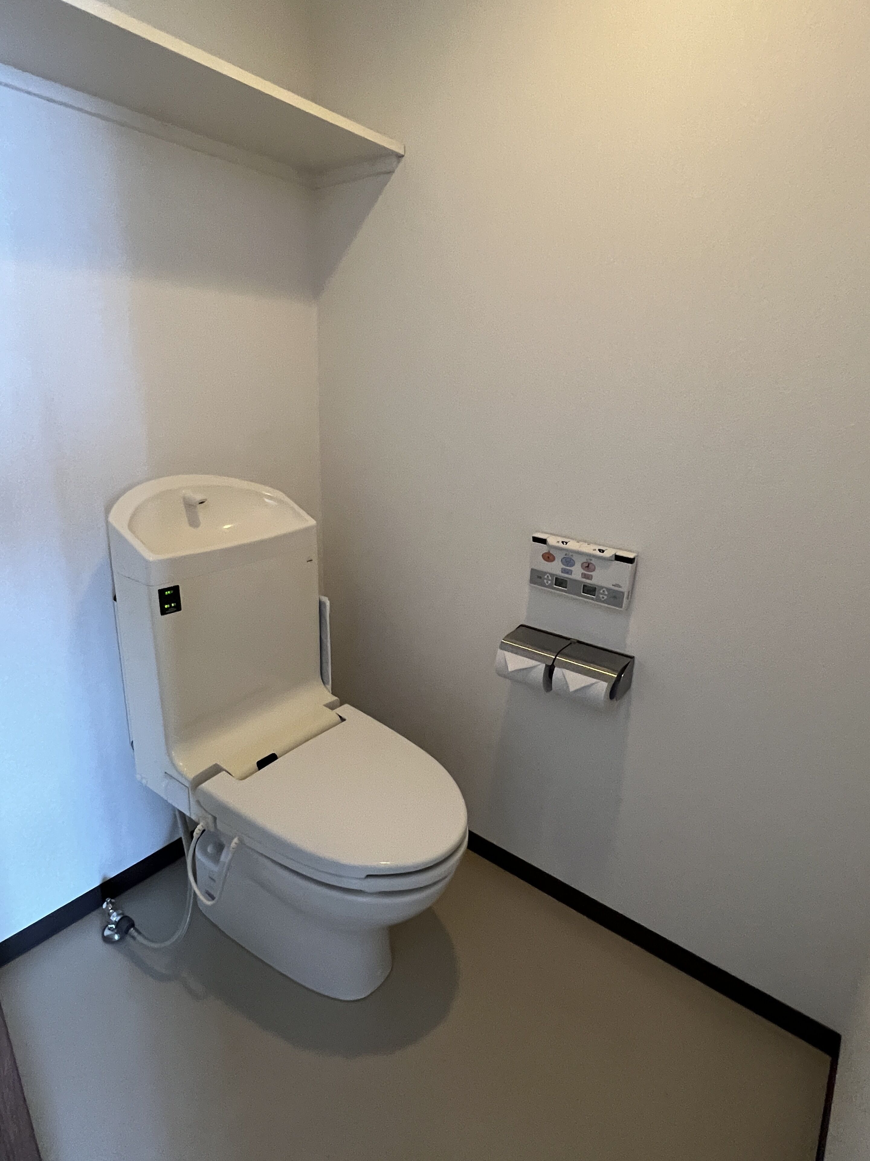 A white toilet with a control panel on the wall, a shelf above it, and a black floor border.