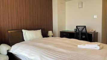 A hotel room with a bed, a desk, a chair, a lamp, and an air conditioning unit.