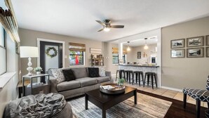A living room with a sofa, coffee table, and ottoman, a kitchen in the background, and a ceiling fan. - 3 Bedroom Home / Hot Tub / Firepit / Game room (Fredericksburg)