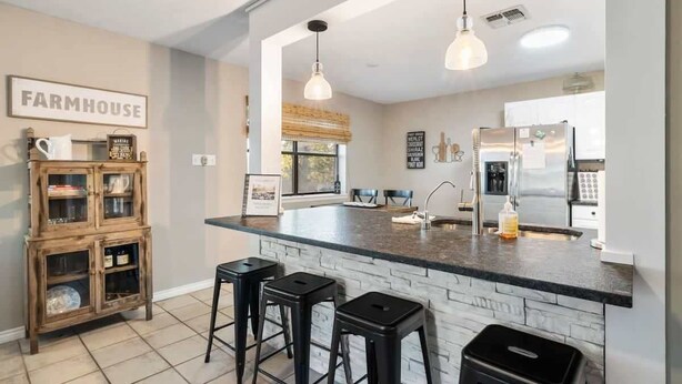 A kitchen with a farmhouse sign, a wooden cabinet, a refrigerator, and two bar stools. - 3 Bedroom Home / Hot Tub / Firepit / Game room (Fredericksburg)