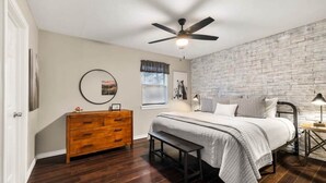 A bedroom with a bed, wooden dresser, bench, ceiling fan, and a brick-patterned wall. - 3 Bedroom Home / Hot Tub / Firepit / Game room (Fredericksburg)