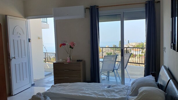 Ein Schlafzimmer mit Bett, Kommode, einer Kommode mit Blumenvase sowie einem Balkon mit Stuhl und Blick aufs Meer.
