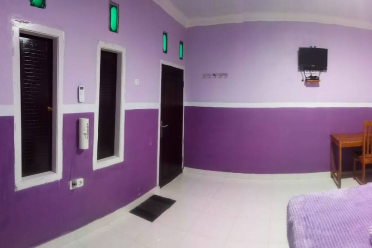Ein Zimmer mit violetten Wänden, einem Bett mit violetter Decke, einem Holzstuhl, einem kleinen Tisch und einem an der Wand befestigten Fernseher.