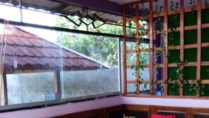 A room with a wooden bench seating area, a small table with a decorative item, and a view of a house through a large window. - Homestay Sukses Tambora (Pekat)