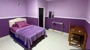 A bedroom with purple walls, a bed with a purple bedspread, a wooden nightstand, a small table, and a mirror on the wall. - Homestay Sukses Tambora (Pekat)