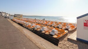 A row of orange and white paddle boats lined up on a beach, with a clear sky and calm sea in the background. - The Esplanade (Weymouth)