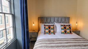 A hotel room with a large window, a bed with a striped blanket, two decorative pillows, and two bedside lamps. - Book House (Whitby)