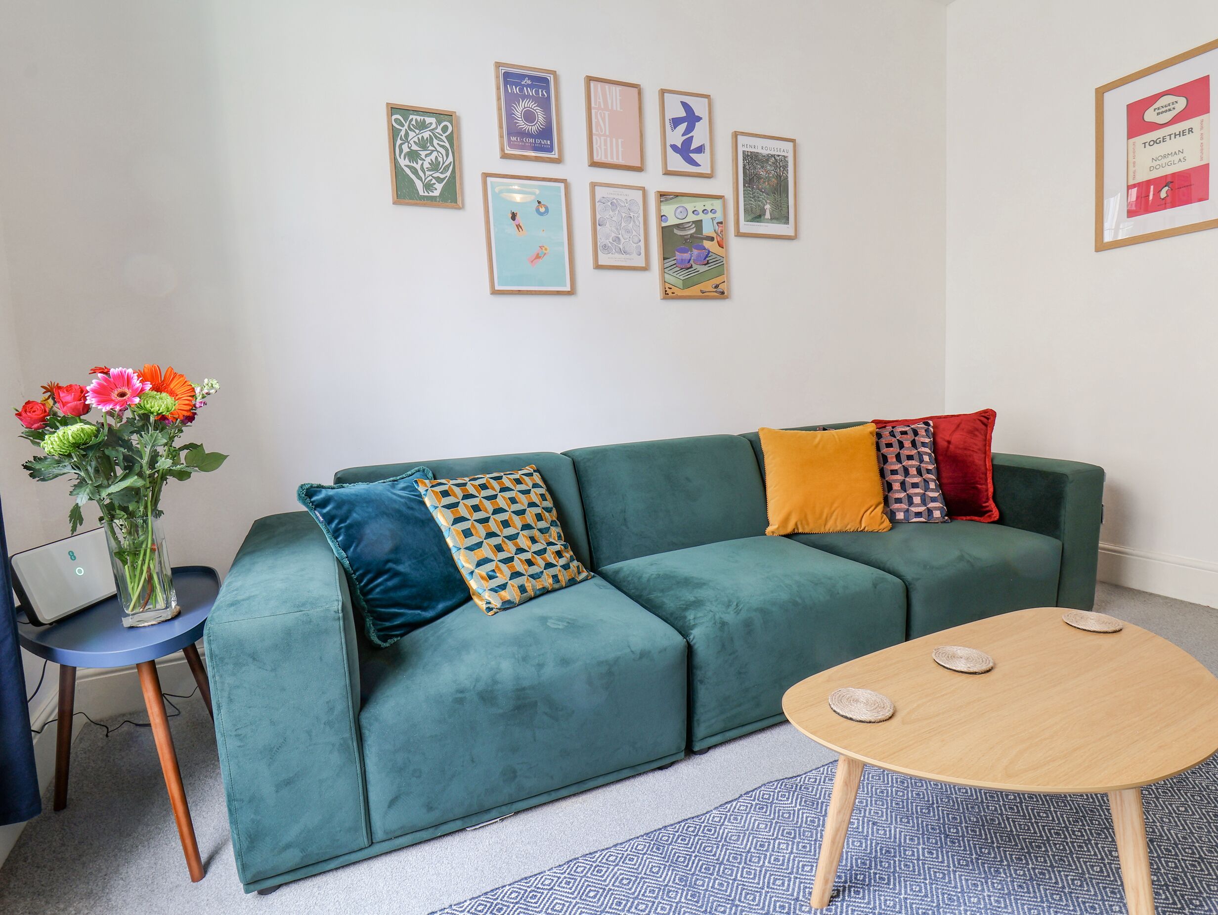 Una sala de estar con un sofá color turquesa, una mesa de centro de madera y un tapete estampado. Hay obras de arte enmarcadas en la pared y un jarrón con flores sobre una mesita.