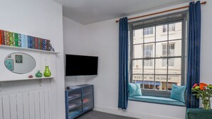 A living room with a television, bookshelves, a window seat with cushions, and a view of a building with signage. - Book House (Whitby)