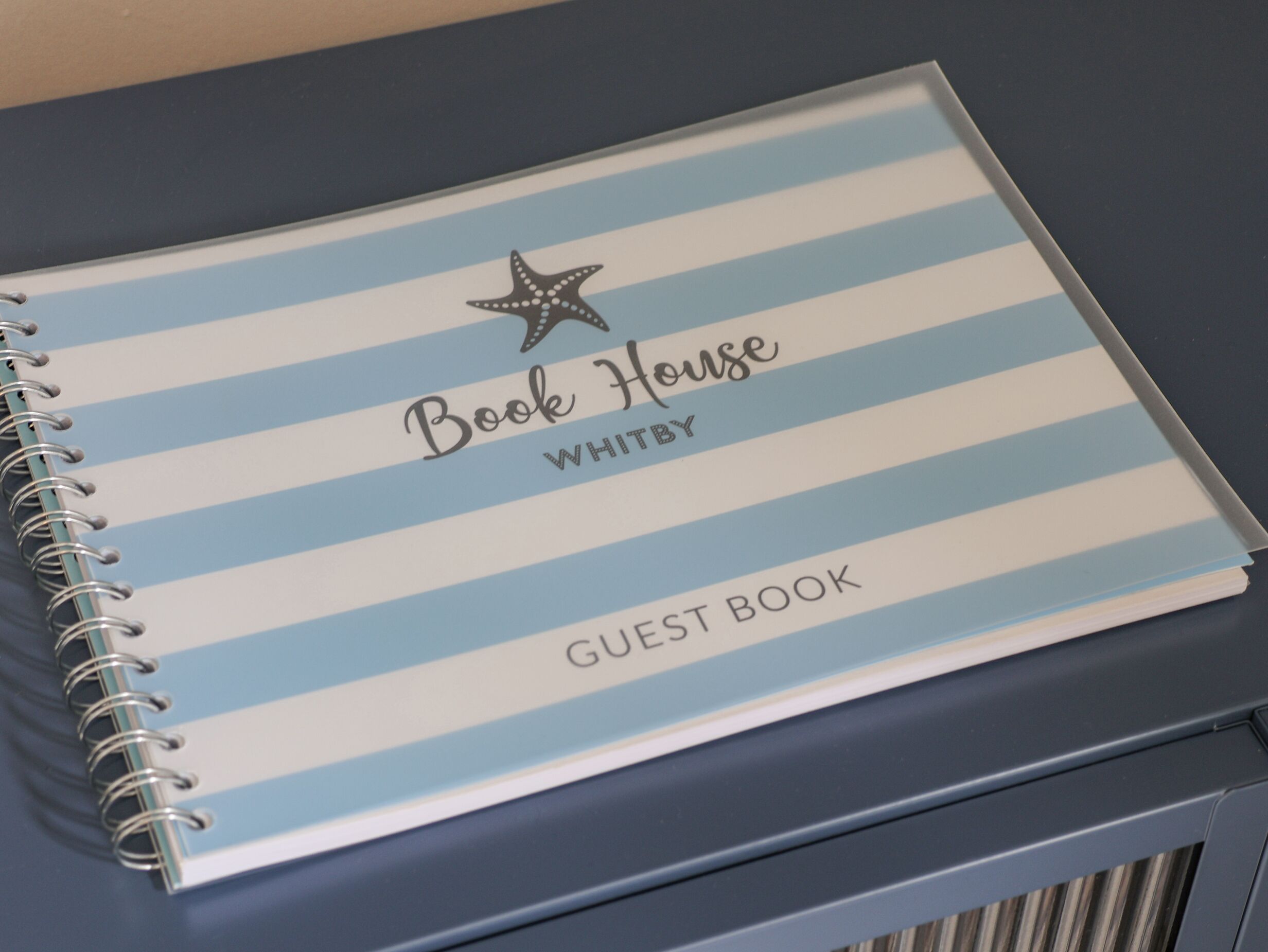 Ein spiraleingebundenes Notizbuch mit blau-weiß gestreiftem Einband, beschriftet mit 'Book House WHITBY GUEST BOOK'.