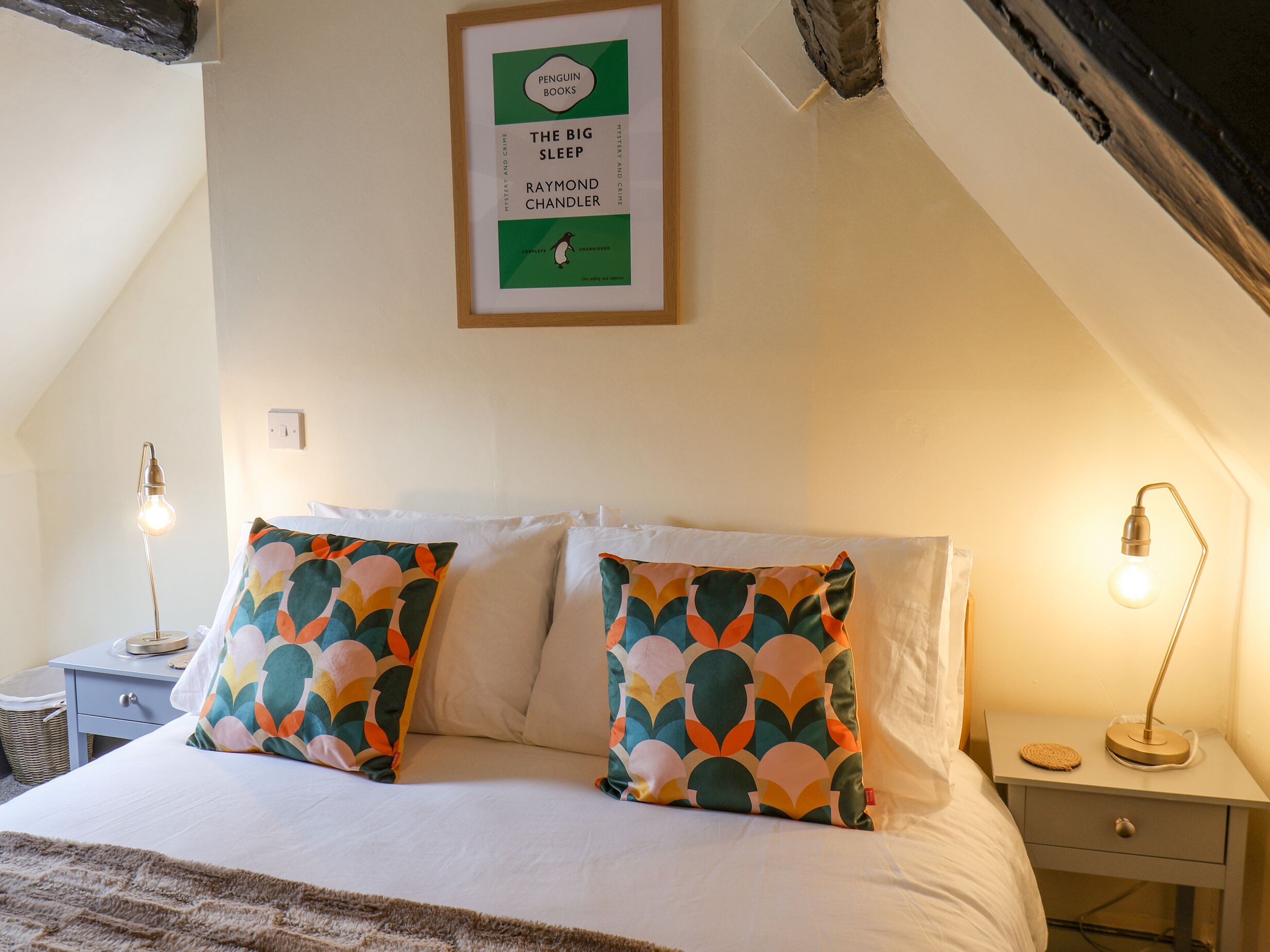 A bedroom with a bed, pillows, a framed book on the wall, bedside tables with lamps, and a small drawer.