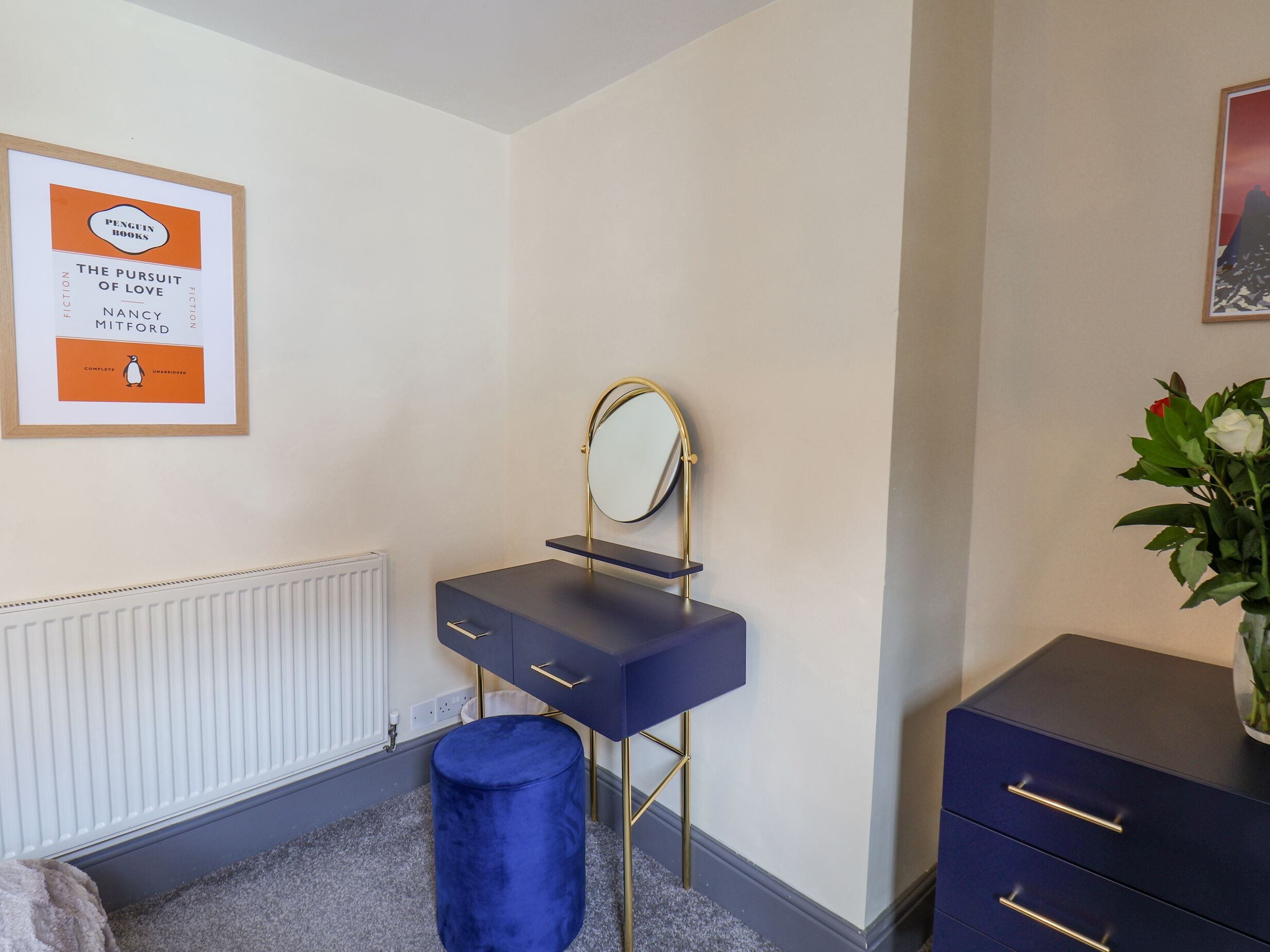 A bedroom with a dark blue vanity, a gold-framed mirror, a blue stool, a radiator, and a framed book on the wall.