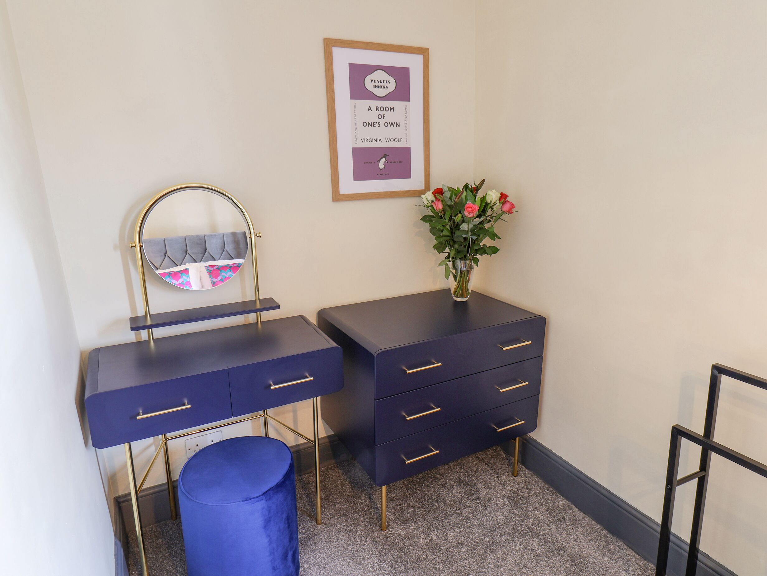 A room with a dark blue vanity, a round mirror, a vase with flowers, and a framed book on the wall.