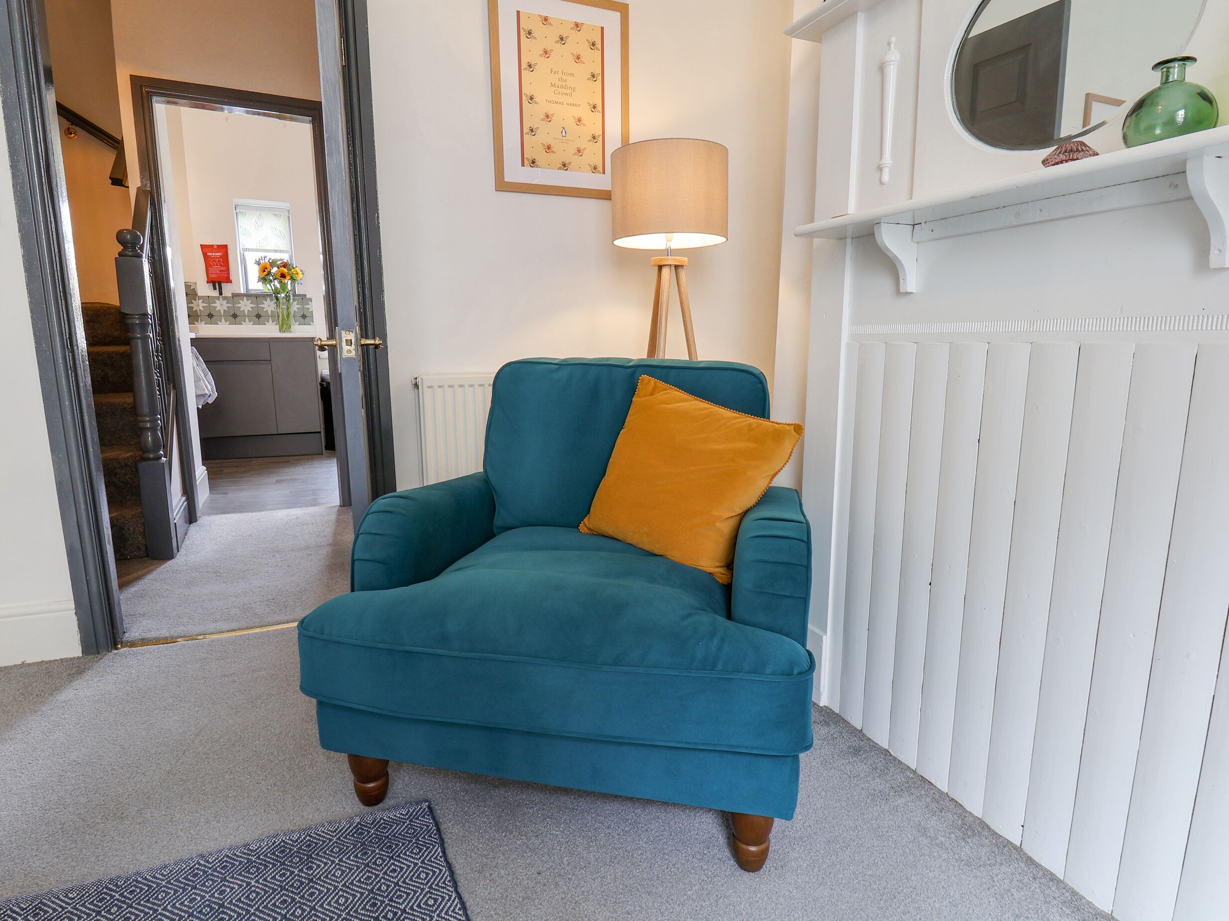 A teal armchair with a mustard-colored pillow in a room with a white fireplace, a framed picture on the wall, and a kitchen visible through an open door.
