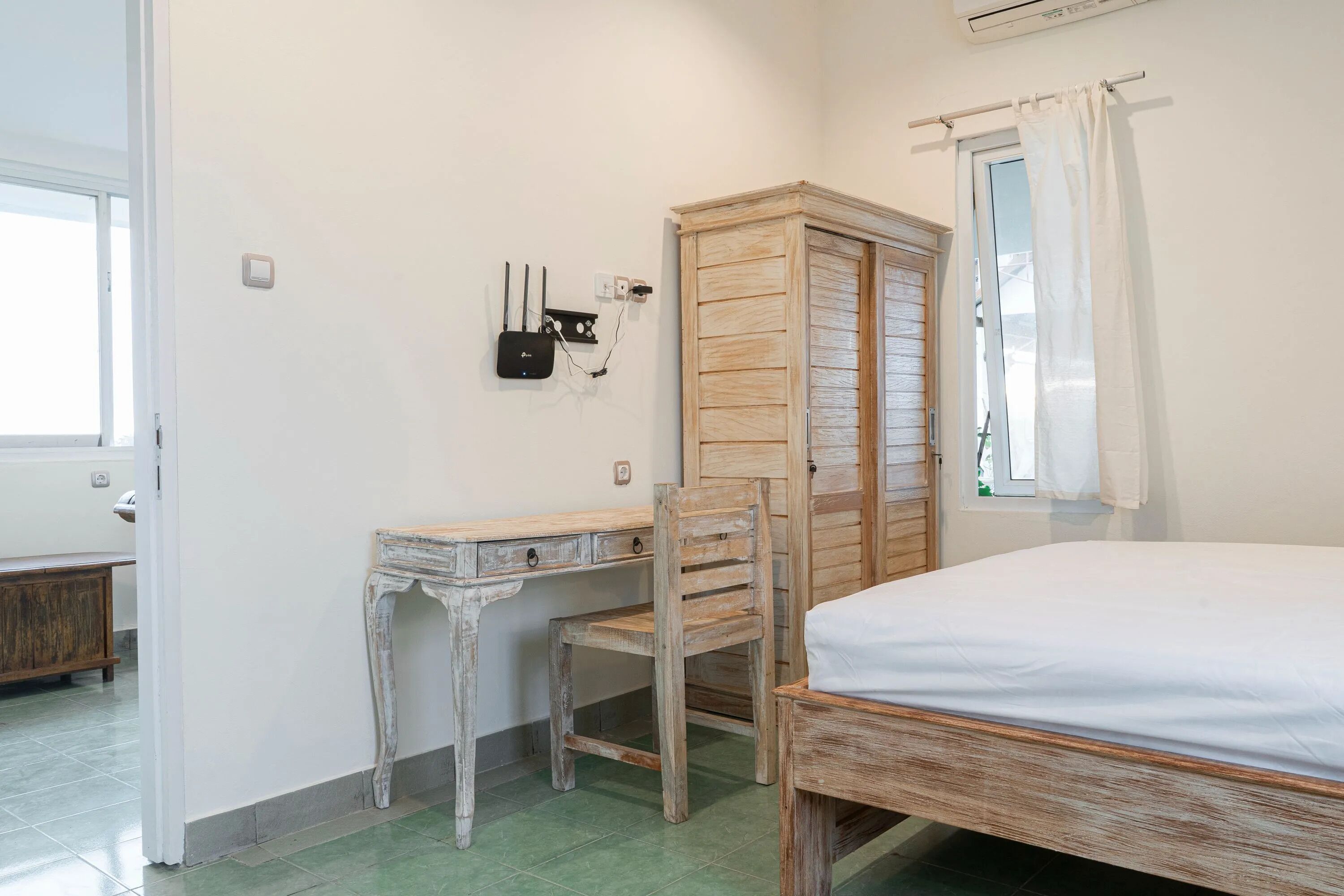 A bedroom with a wooden bed, a desk, and a chair. There is a wardrobe, a window with curtains, and an air conditioning unit on the wall.