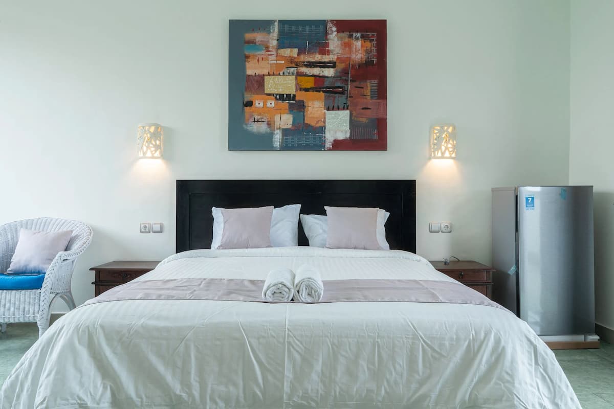 A hotel room with a large bed, two bedside tables, a white chair, a wall-mounted artwork, and a refrigerator.
