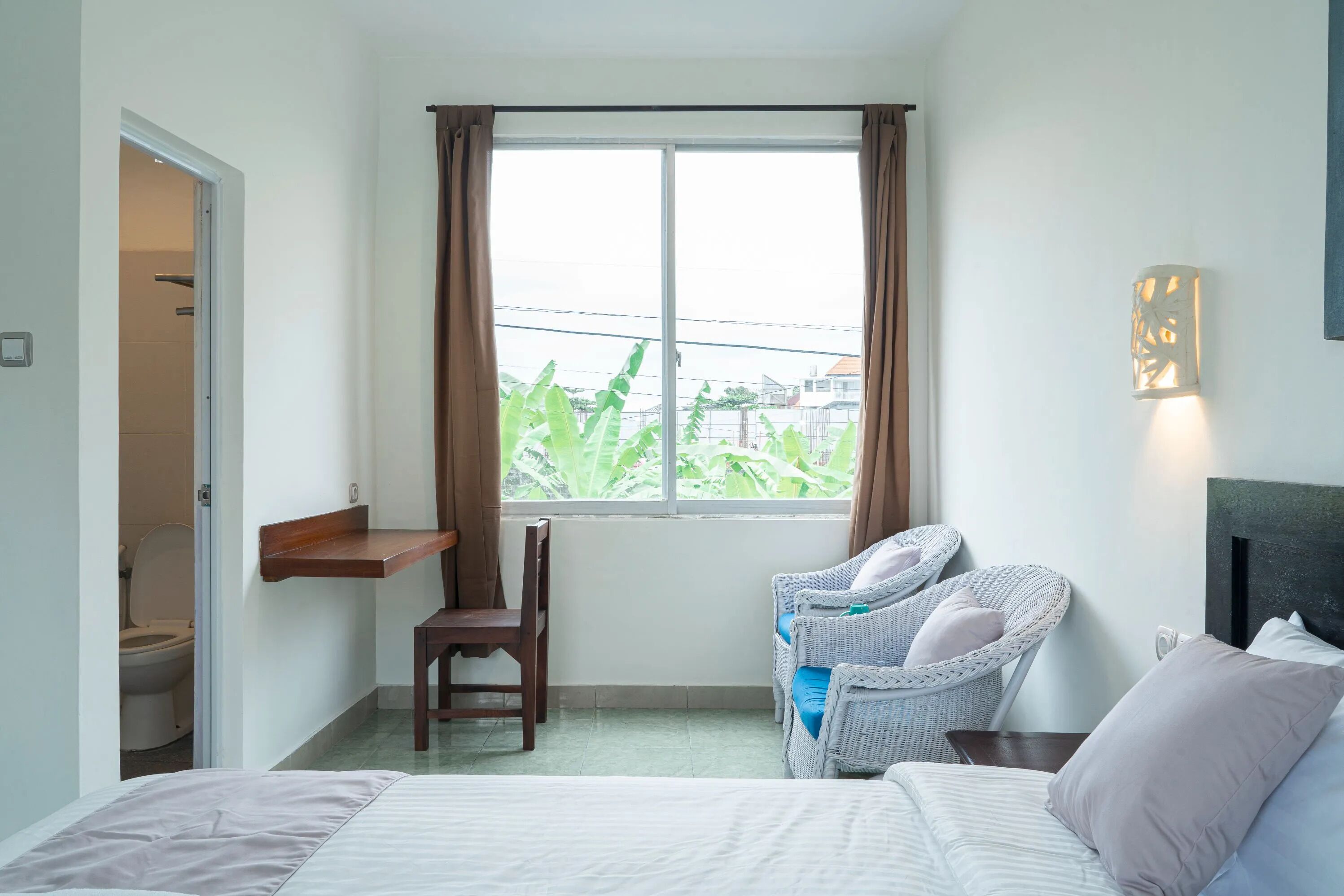 A hotel room with a bed, a chair, a desk, a window with curtains, and a wall-mounted light fixture.
