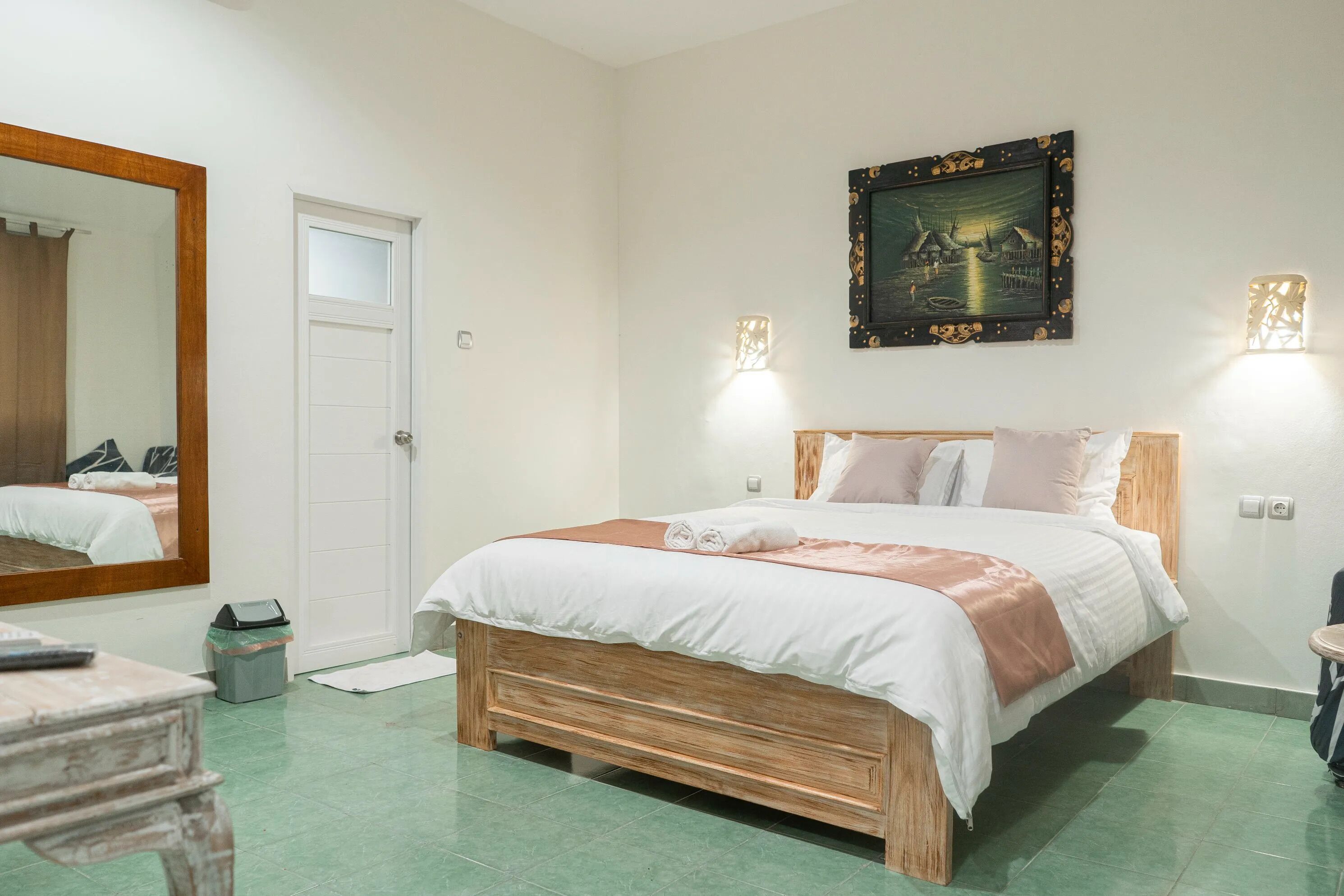 A bedroom with a wooden bed, white bedding, a large mirror, a framed picture, and two wall-mounted lights.