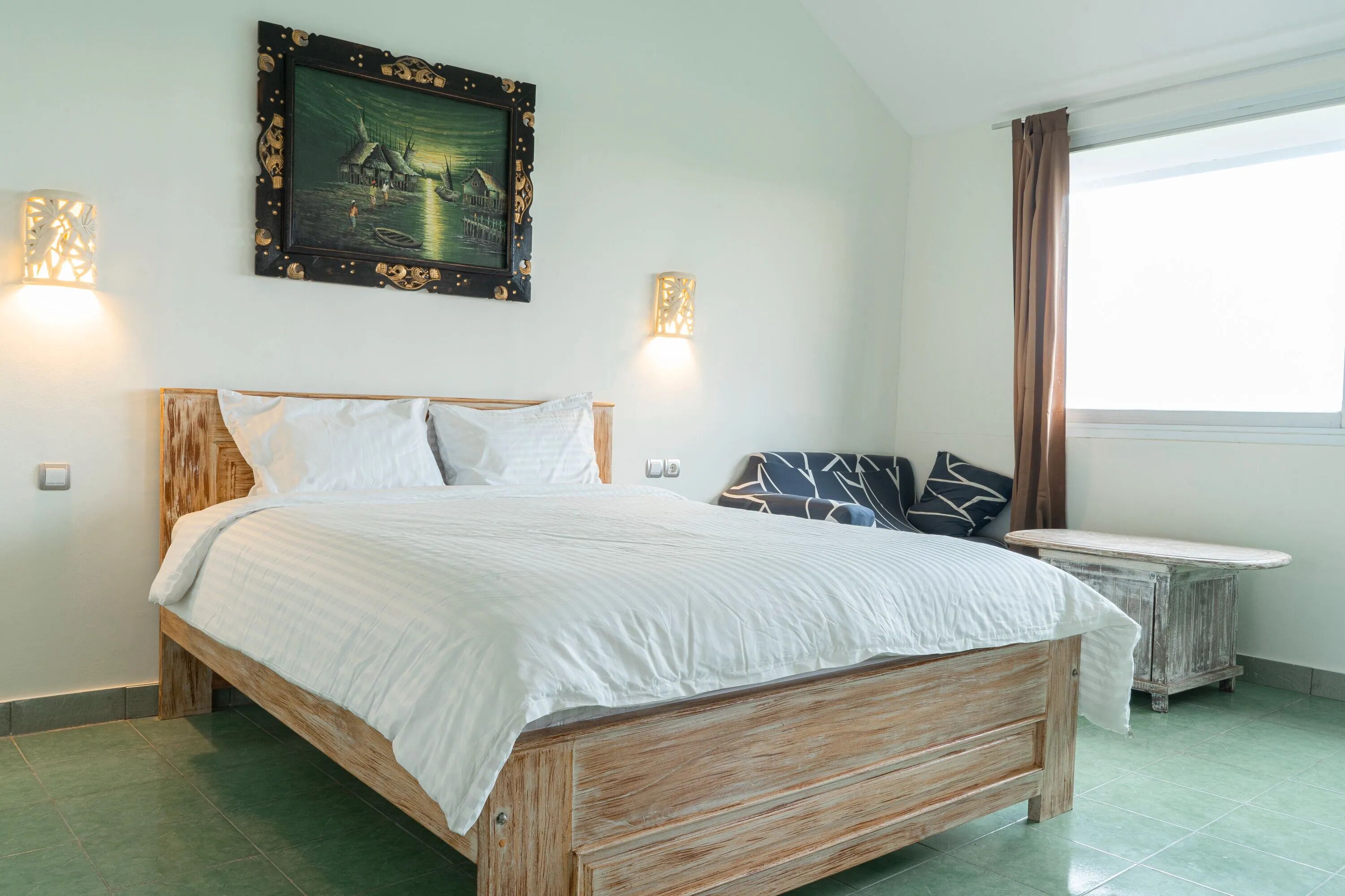 Una camera da letto con un letto in legno, biancheria bianca, un quadro appeso al muro, due applique e una finestra con tende.