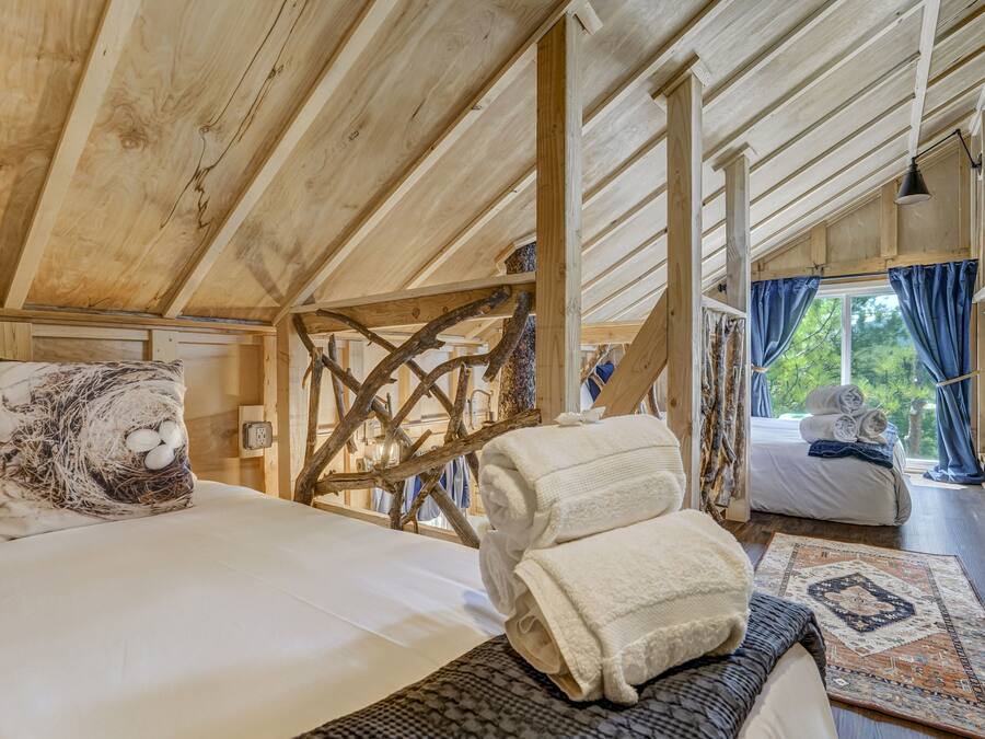 Ein gemütliches Schlafzimmer in einer Holzhütte mit einem Schlafplatz in der Mansarde, einer rustikalen Holzleiter und Blick ins Freie durch ein Fenster mit blauen Vorhängen.
