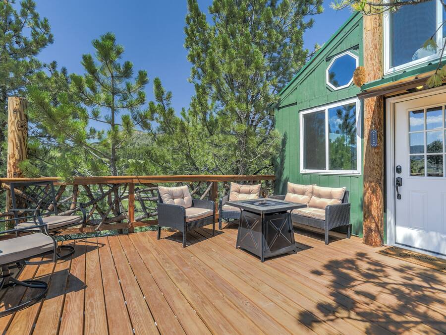 A wooden deck with outdoor furniture, a green building with a white door, and surrounding pine trees.